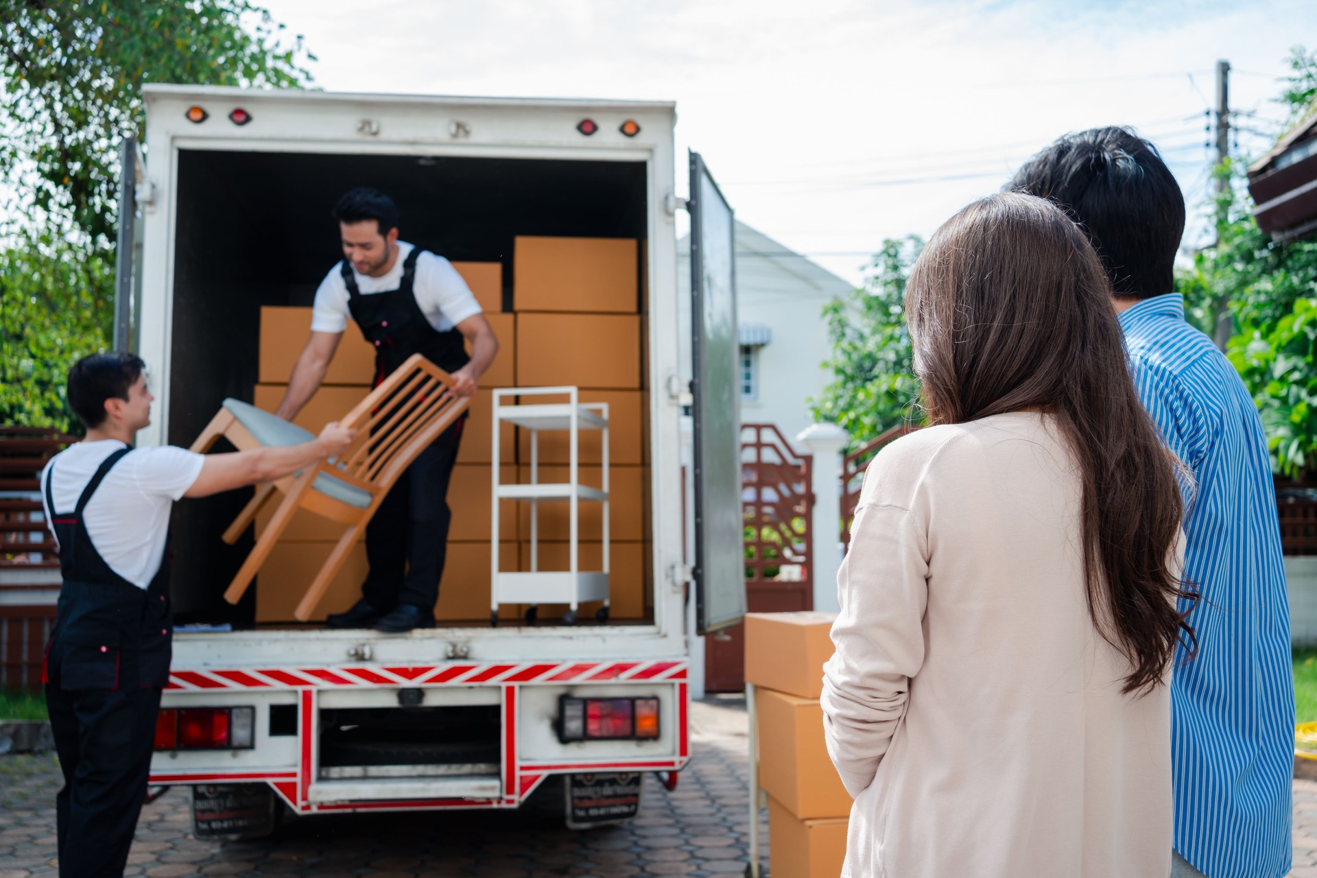 Asian Couple check while unloading boxes and furniture from a pickup truck to a new house with service cargo two men movers worker in uniform lifting boxes. concept of Home moving and delivery.